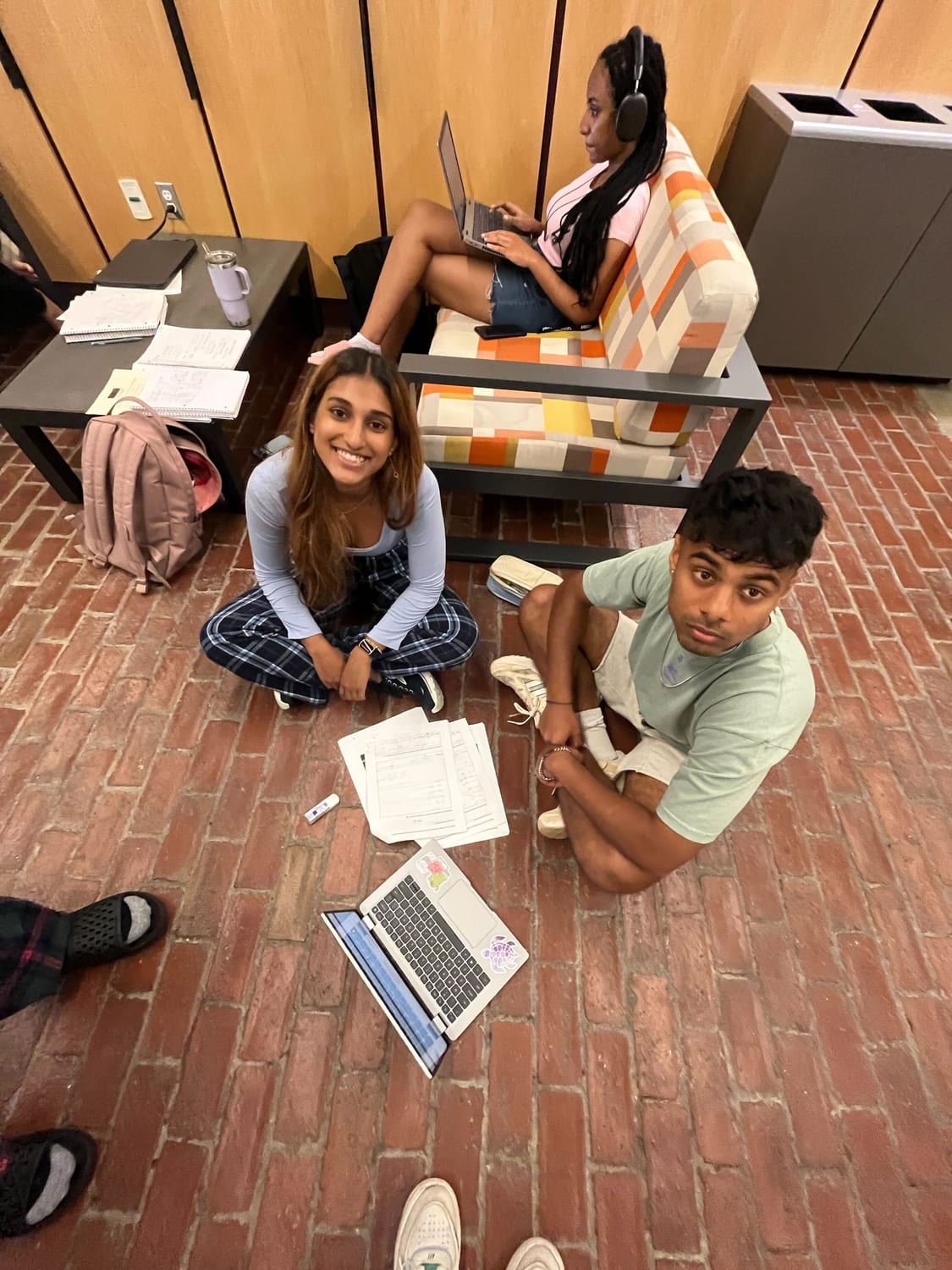 Ava and two classmates sitting on the brick floor of a dorm lounge late at night, laptops and problem sets spread around them.