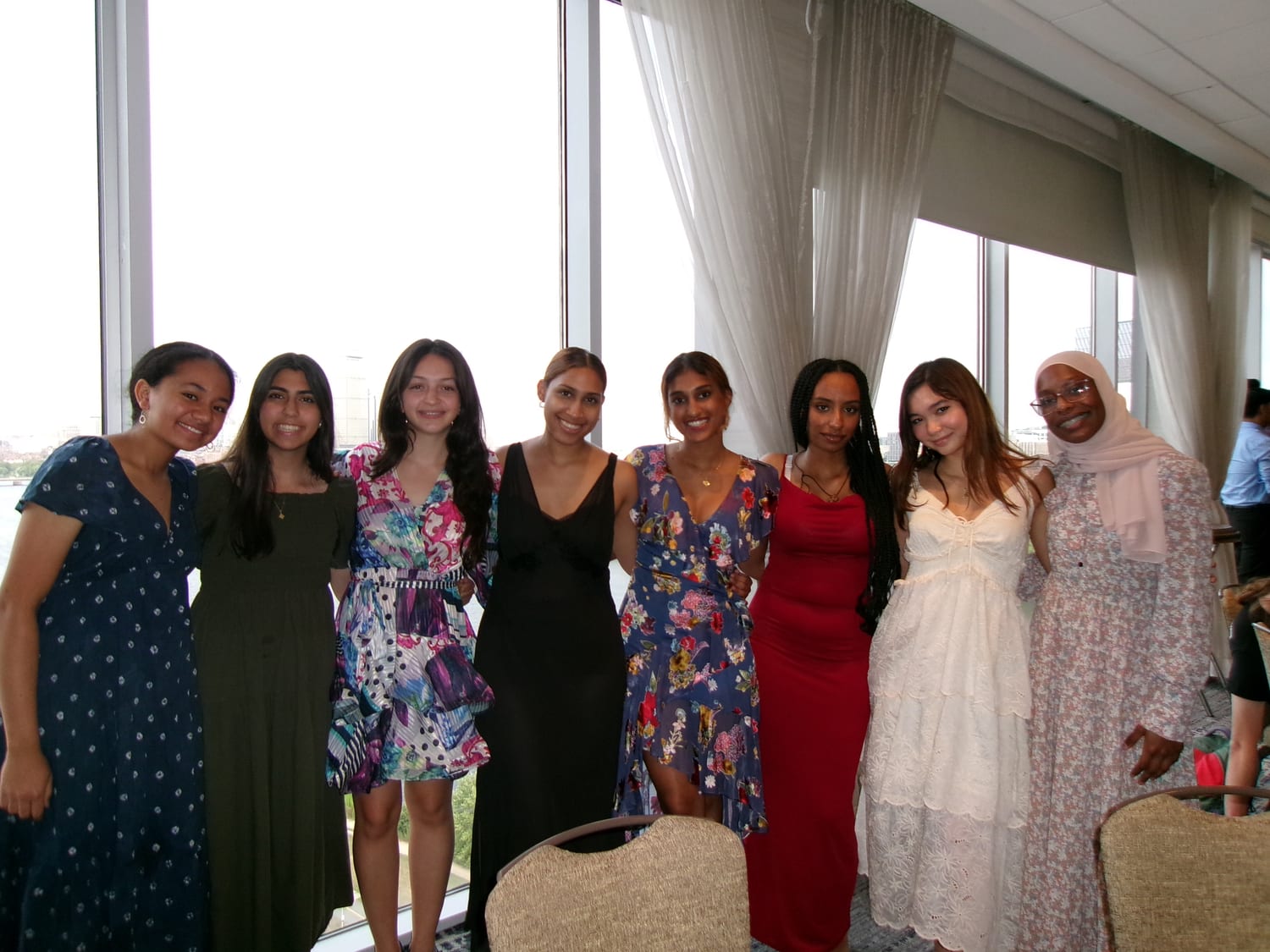 Eight students in formalwear arm in arm in front of a window overlooking the Charles at the closing banquet.