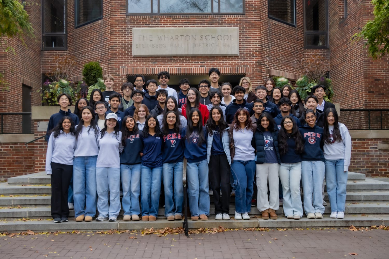 The M&T cohort — around forty-five students, most in navy Penn sweatshirts and jeans — banked up the steps of the Wharton School on an autumn morning, the carved lettering “The Wharton School — Steinberg Hall · Dietrich Hall” above them and fallen leaves on the brick.
