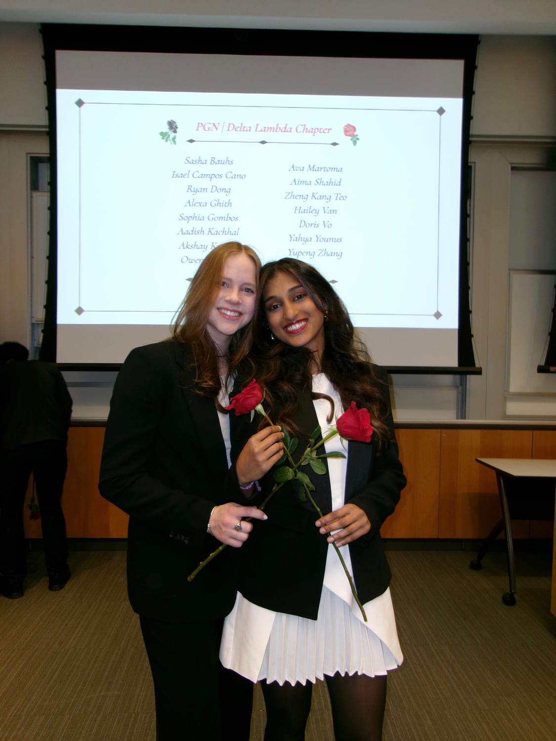 Ava and a fellow new member in black blazers, each holding a long-stemmed red rose, in front of a projected slide headed “PGN | Delta Lambda Chapter” listing the new-member class with Ava Martoma among the names.