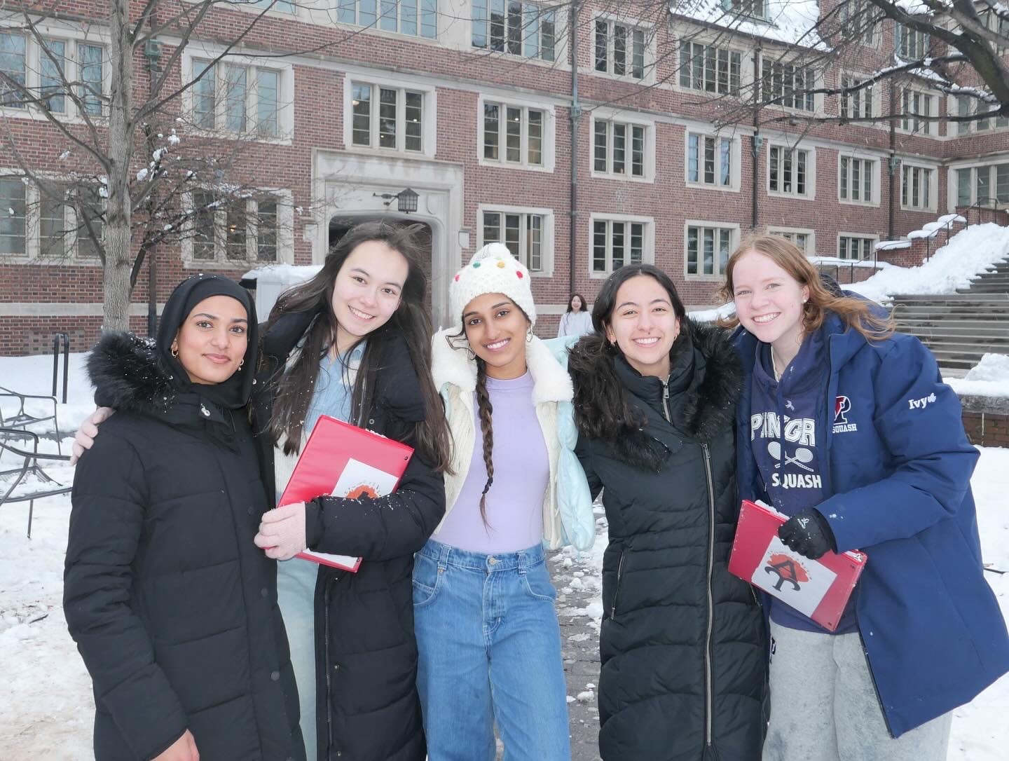 Five members in heavy winter coats, one in a navy Penn squash jacket, arms around each other in the snow outside a brick residence hall, two of them carrying red binders.