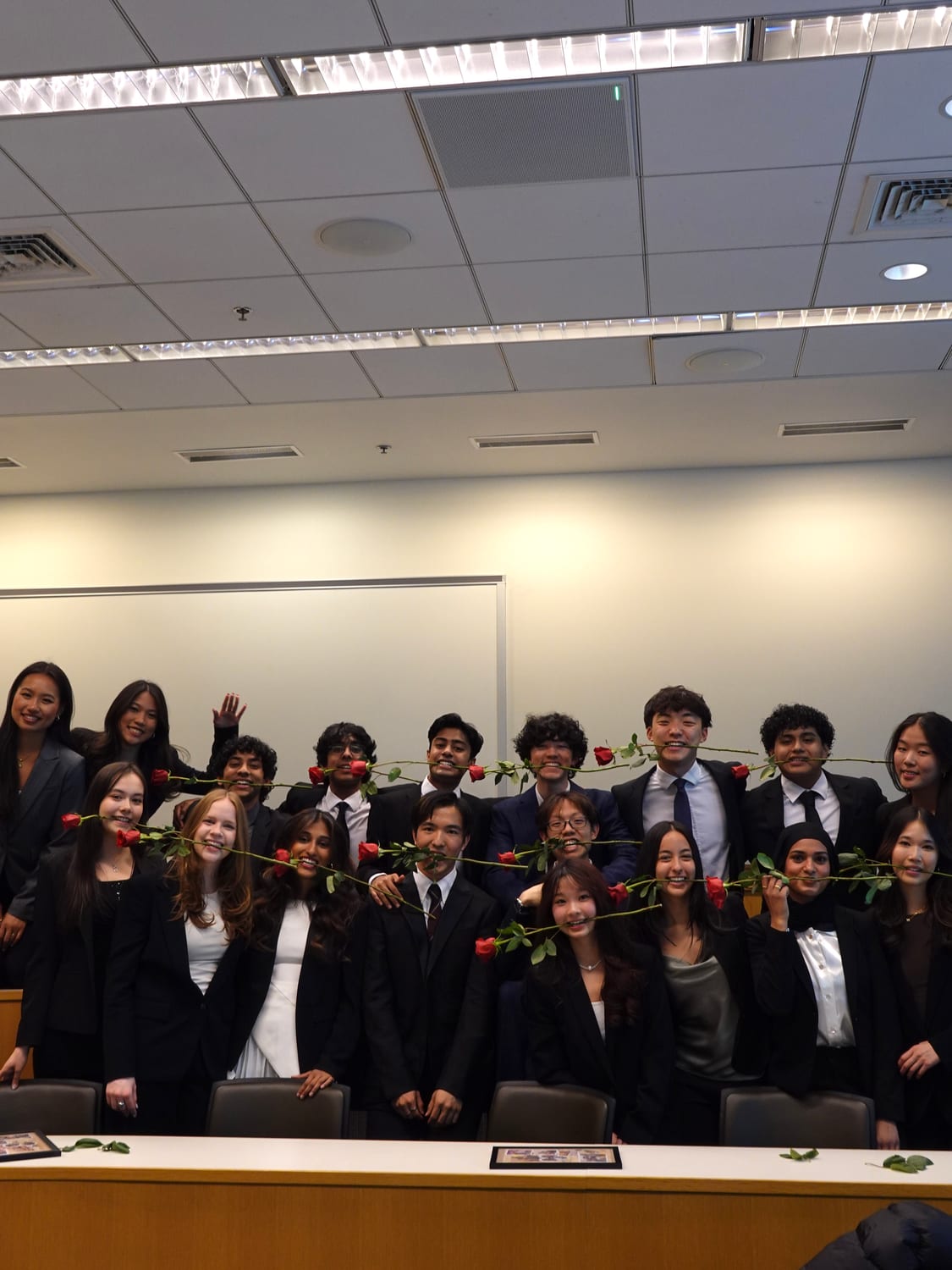 Around twenty members in business formal behind a long classroom desk, passing red roses down the row and holding them in their teeth, loose petals and leaves scattered across the desk.