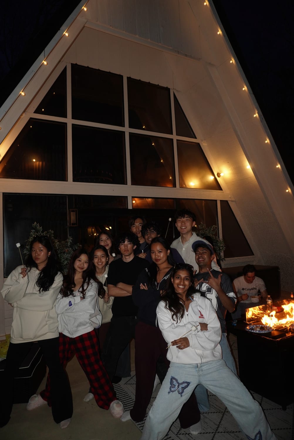 A dozen members in hoodies and pyjama trousers posing with marshmallow skewers outside a string-lit A-frame house at night, a fire pit burning off to one side.