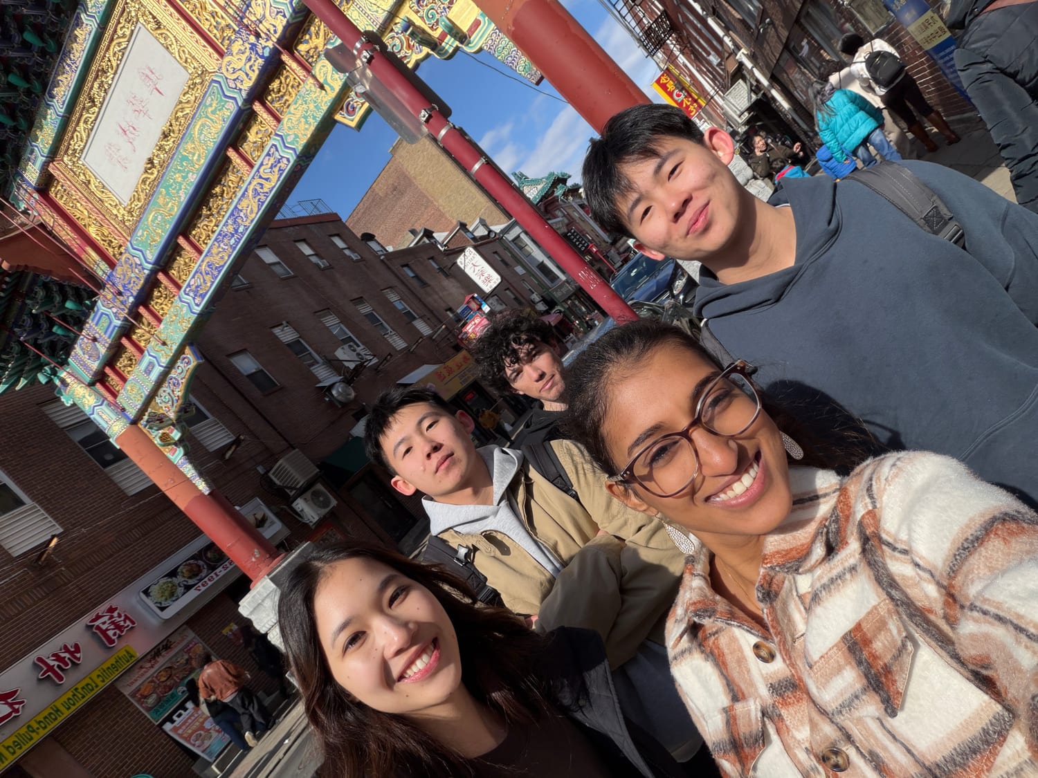 A tilted five-person selfie beneath a red, green and gold Chinatown gate, hand-pulled-noodle shopfronts running down the street behind.