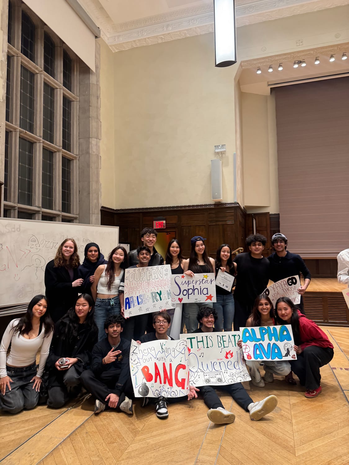 Around eighteen members on the parquet floor of a wood-panelled hall holding hand-made posters — “Alpha Ava”, “Superstar Sophia”, “Our fav house dancers Aadish + Ryan”, “Zheng Kang out with a BANG” — with “PGN” scrawled on the whiteboard behind them.