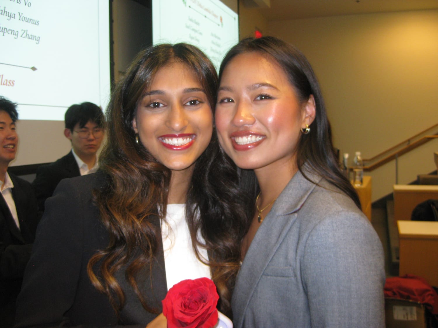 Ava and another new member cheek to cheek in front of the projector screen, a red rose held up between them and the last few names of the class list still visible behind.