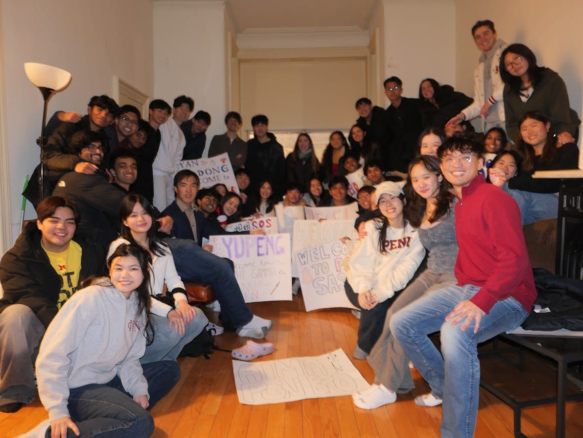 About forty members packed into a chapter-house room at night, sitting on the floor, on couches and perched on a table, welcome posters held up through the crowd and a grey ΦΓΝ crewneck in the front row.