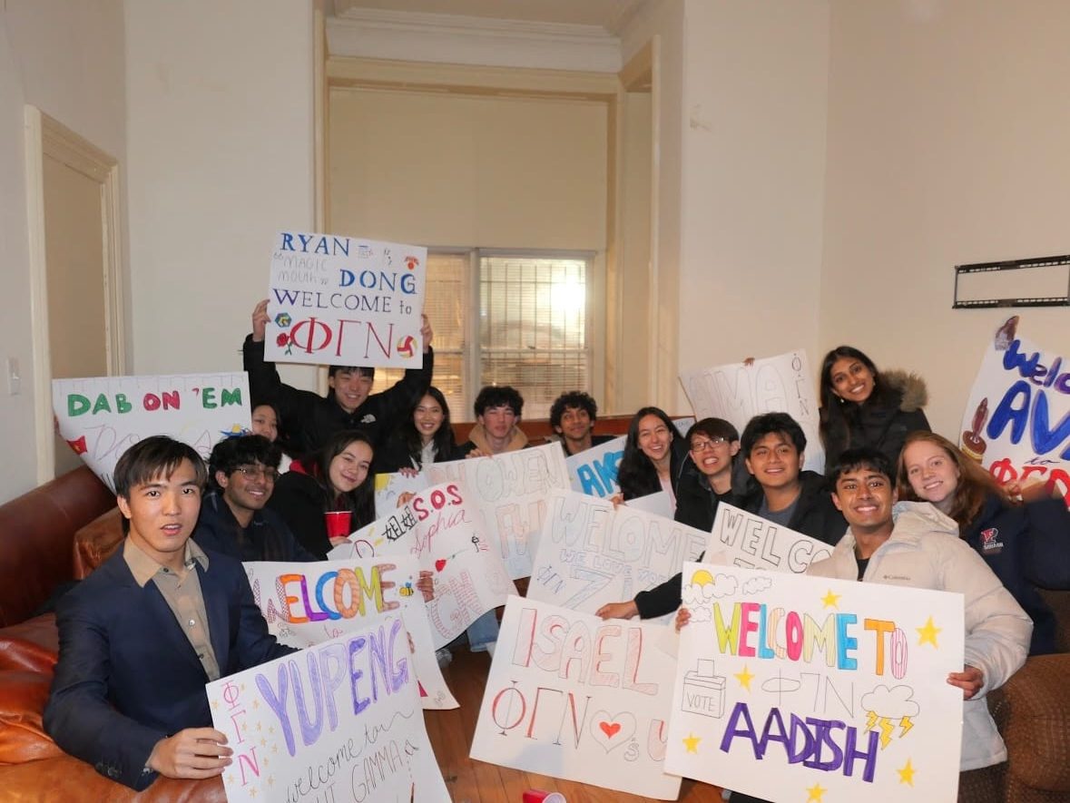 A dozen members crowded onto a leather couch in a chapter-house living room, each holding up a hand-lettered poster — “Ryan Dong, welcome to ΦΓΝ”, “Welcome to ΦΓΝ Aadish”, “Isael”, “Yupeng”, “Dab on 'em” — with one reading “Welcome Ava to ΦΓΝ” at the edge of the frame.