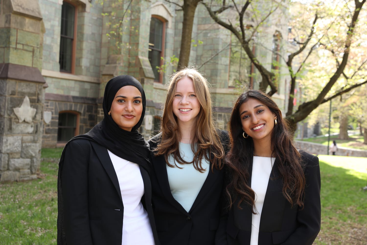 Ava and two fellow members in black blazers over white, arms around each other on the grass in front of one of Penn’s green serpentine-stone halls, the trees just coming into leaf.