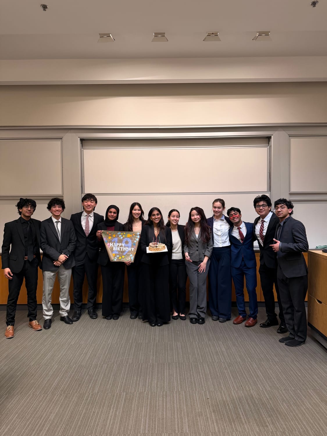 Thirteen members in business formal lined up along a classroom wall; one holds a hand-painted banner reading “Happy Birthday Ava — 19” and Ava, next to her, holds a cake with lit candles.