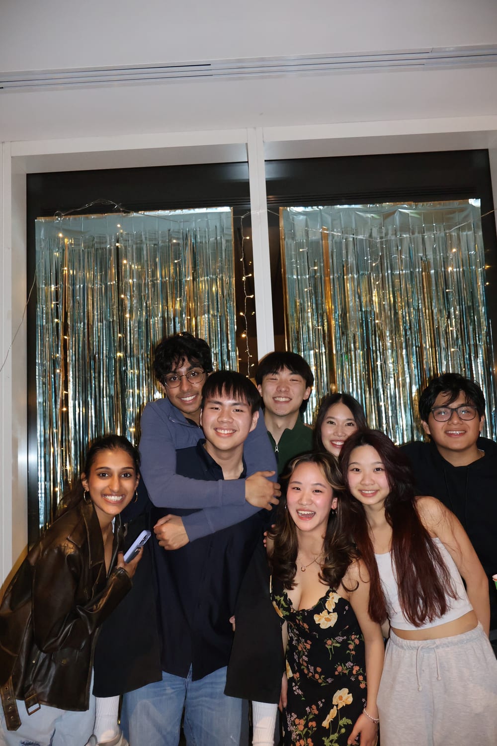 Eight members crowded together in front of a silver tinsel curtain strung with fairy lights at a party.
