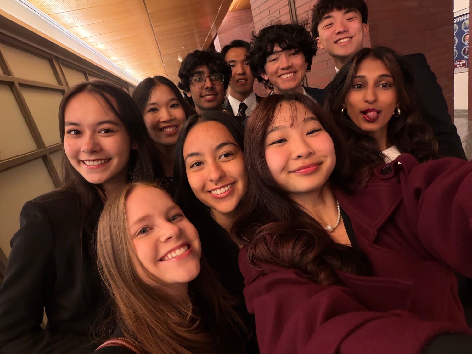 A nine-person selfie in a brick corridor, everyone in suits and blazers, Ava pulling a face at the right-hand edge of the frame.