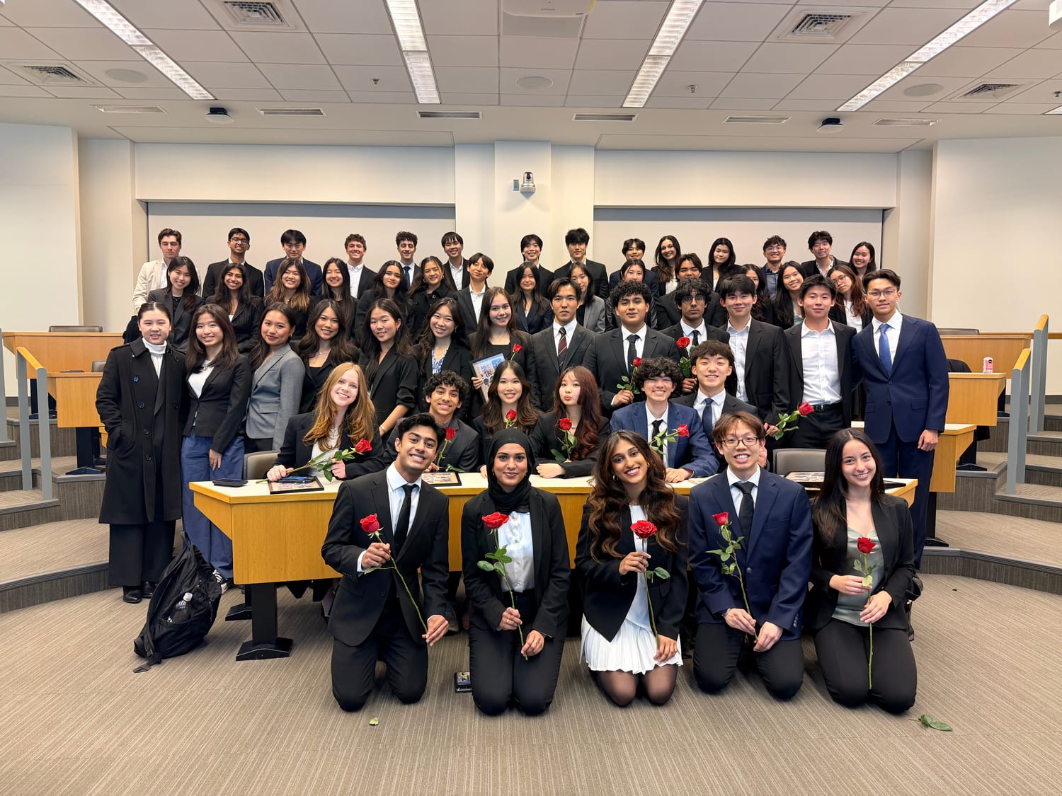 The full chapter — about fifty people in business formal — banked up the tiers of a lecture room, with the new members kneeling along the front holding red roses and framed photographs propped on the desk behind them.