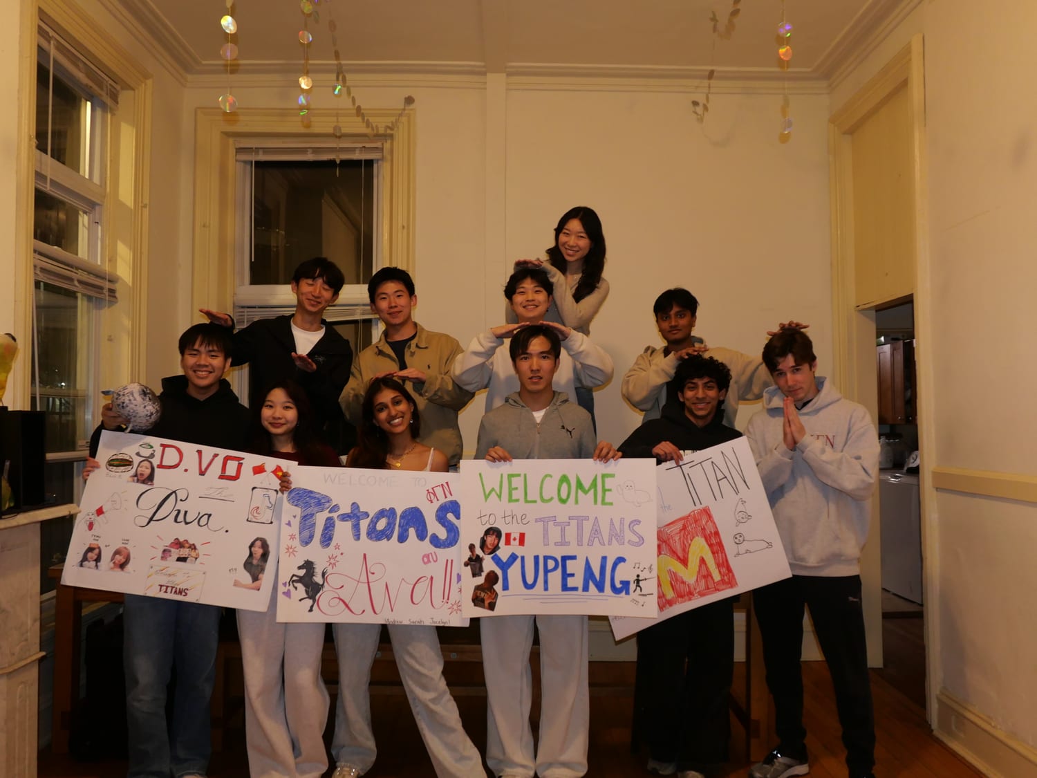 Eleven members in a chapter-house room holding hand-drawn posters that read “Welcome to the Titans” — one for Ava, one for Yupeng, one for D. Vo — while the row behind them make hand-over-head gestures.