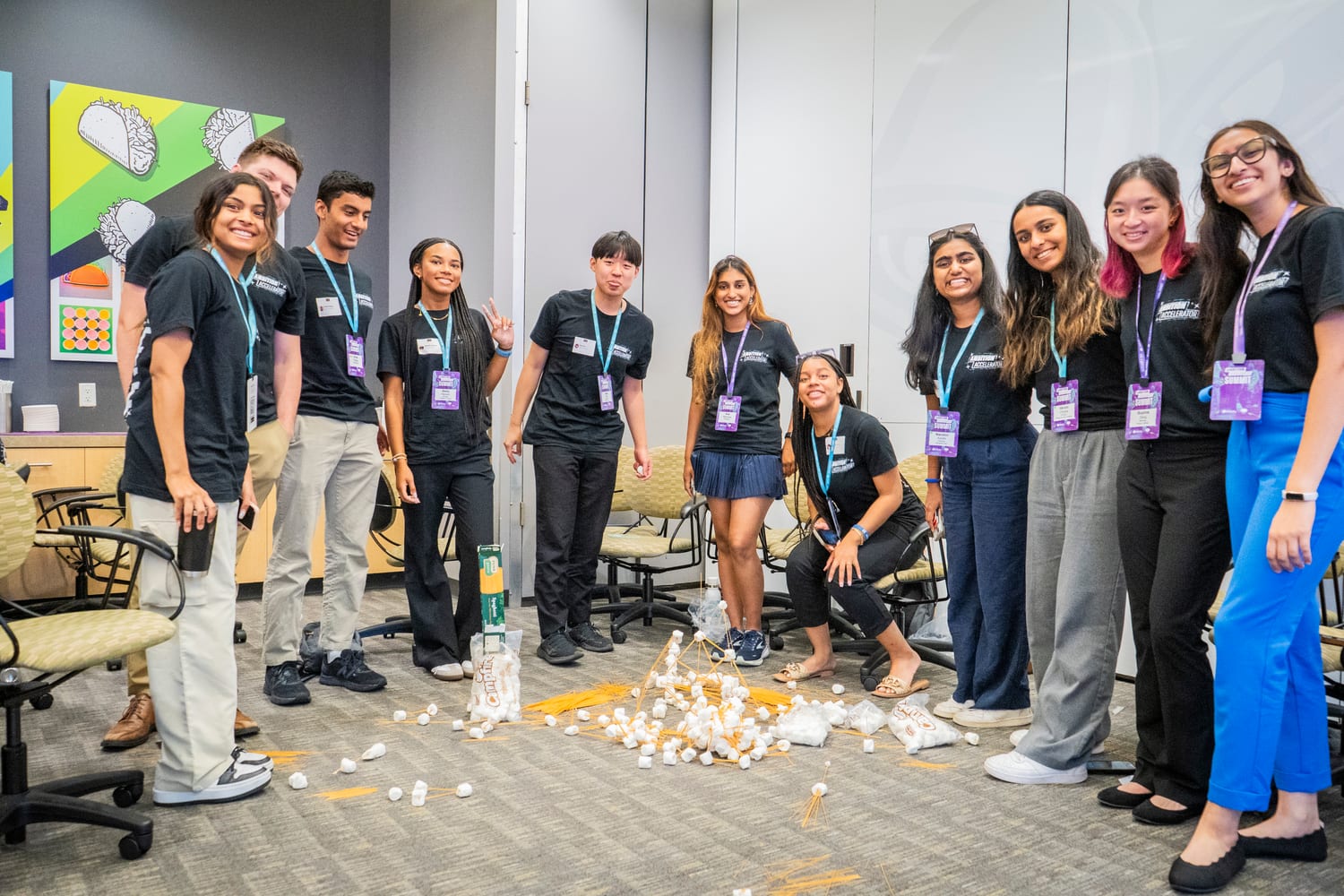 A dozen participants laughing in a conference room around a collapsed tower of spaghetti and marshmallows on the floor, taco artwork on the wall behind them.