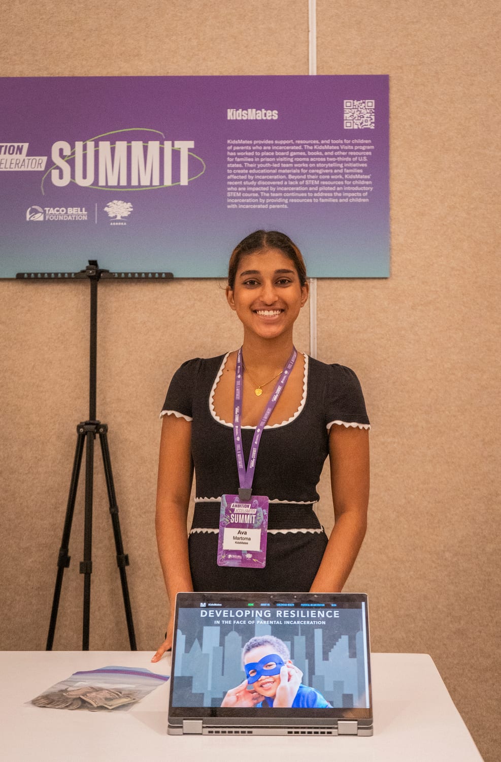 Ava standing behind her KidsMates table at the Ambition Accelerator Summit, a laptop in front of her showing a slide titled “Developing Resilience in the Face of Parental Incarceration”, with a large Taco Bell Foundation and Ashoka summit board describing KidsMates behind her.