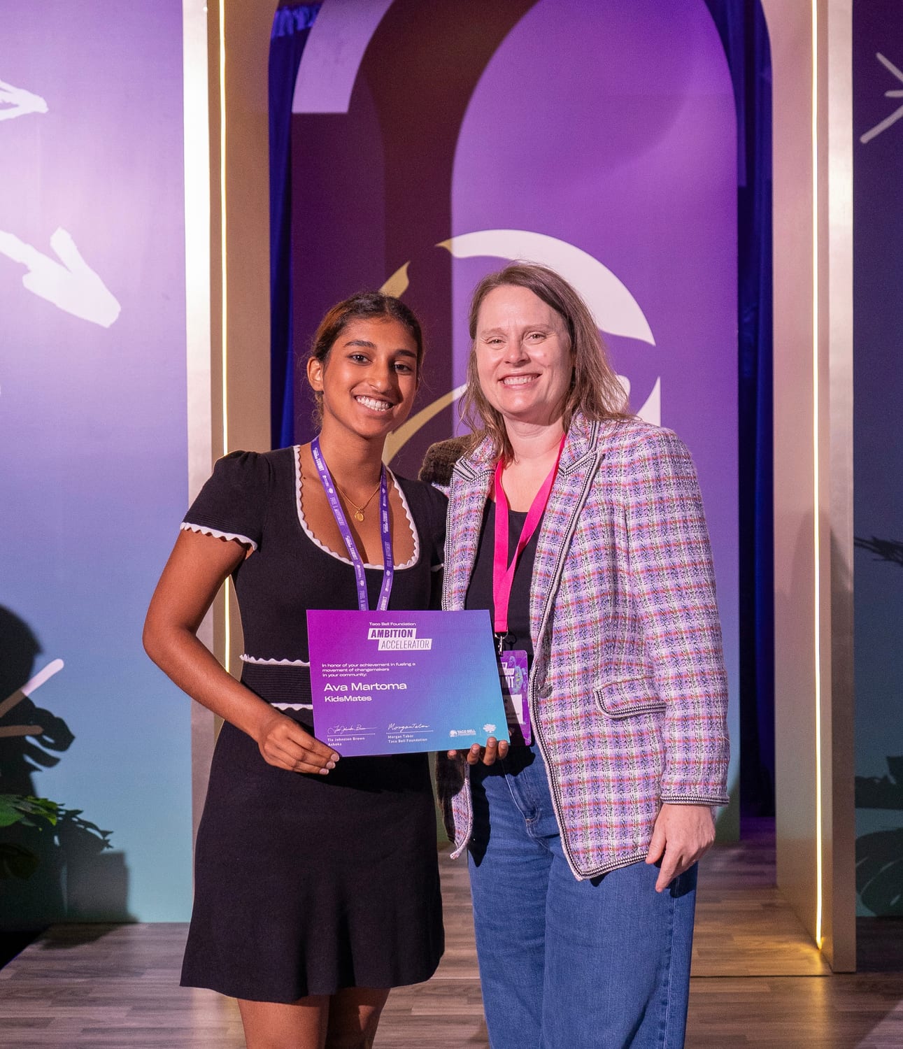 Ava on the summit stage holding her purple Ambition Accelerator certificate, which reads “In honor of your achievement in fueling a movement of changemakers in your community — Ava Martoma, KidsMates”.