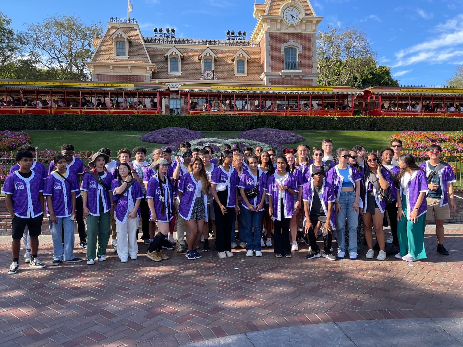 The cohort in purple Taco Bell Foundation jerseys posed in front of the Disneyland Railroad station and the Mickey Mouse flower bed.