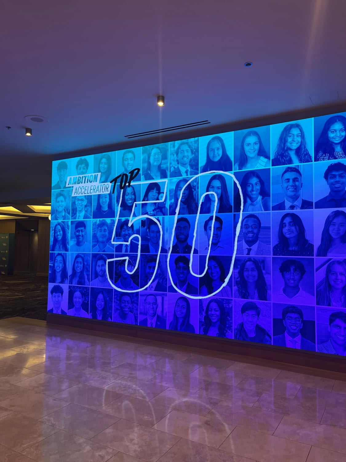 A wall-sized backlit display in the hotel lobby showing a grid of fifty headshots with a hand-drawn “50” across it, captioned “Ambition Accelerator Top 50”.