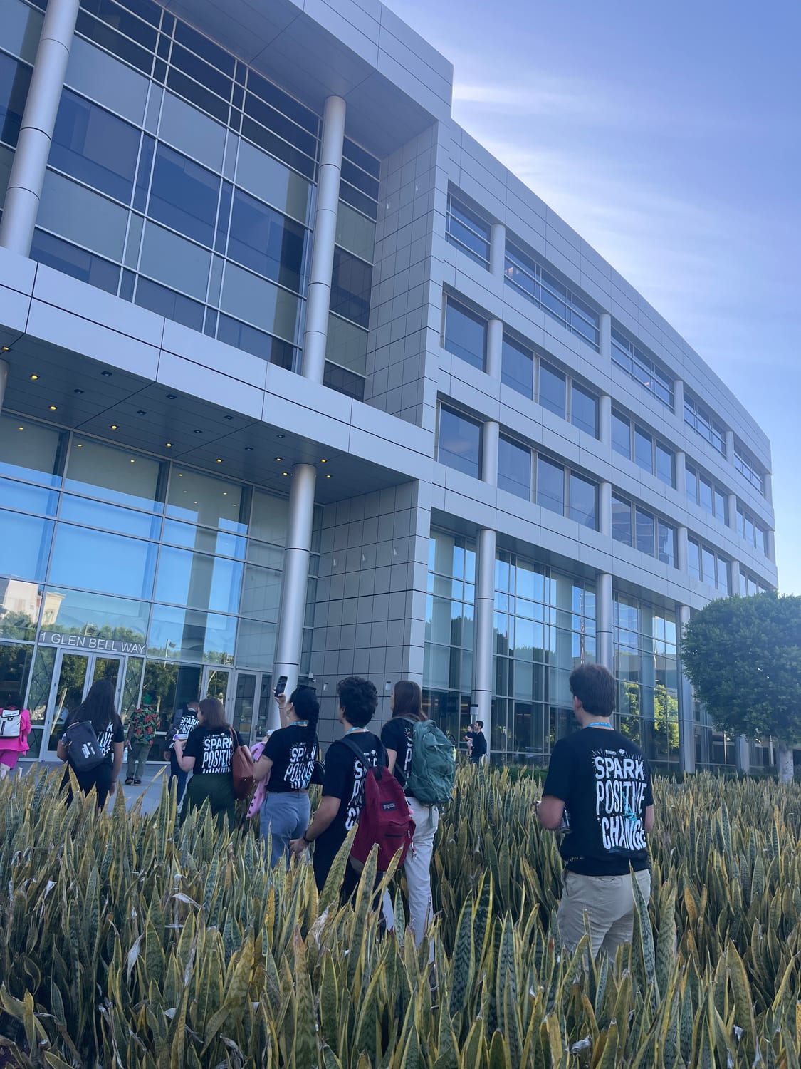Participants in black “Spark Positive Change” T-shirts walking toward the glass doors of Taco Bell headquarters, the entrance lettered “1 Glen Bell Way”.