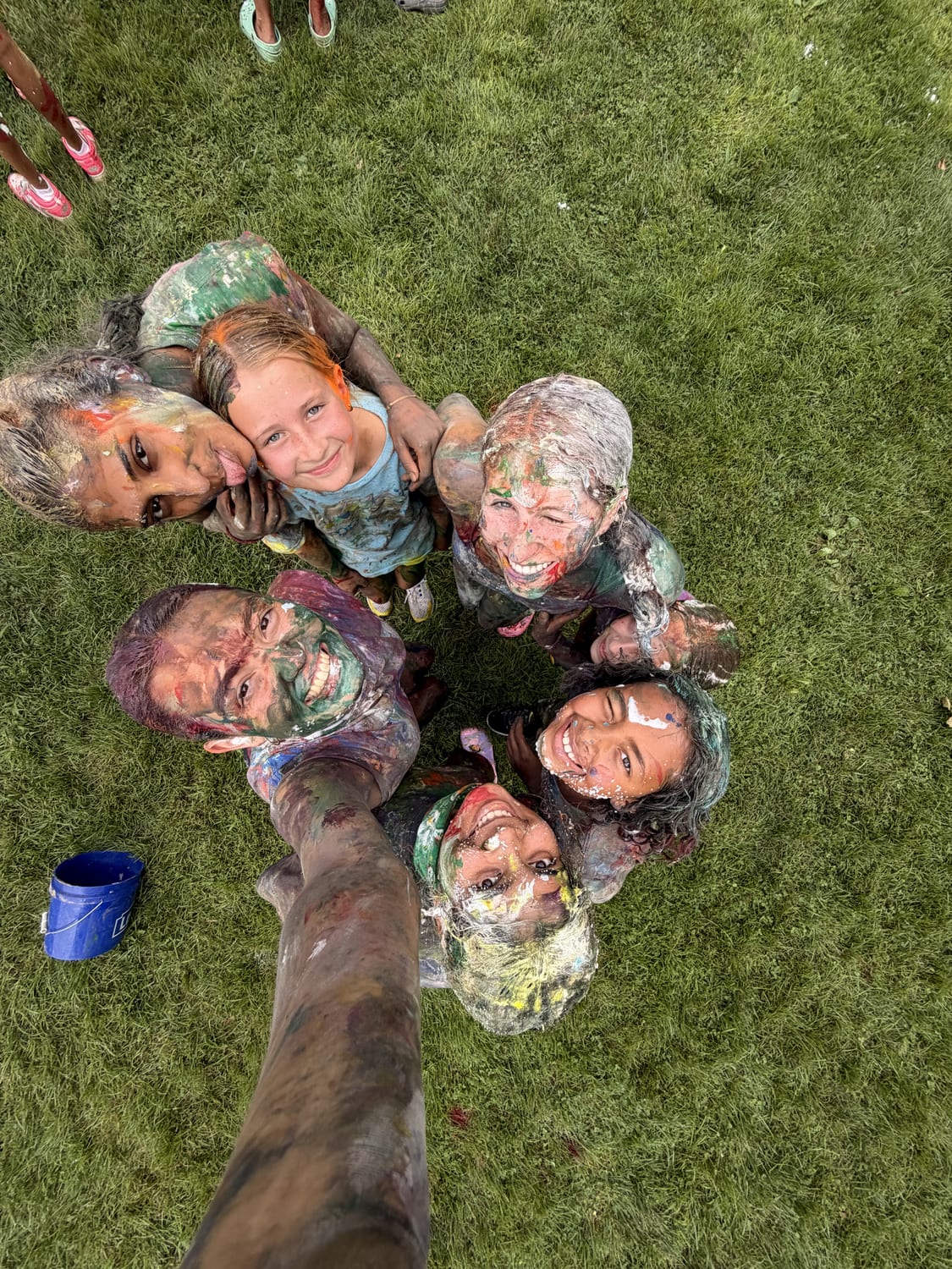 Overhead selfie of a circle of campers and counselors on the grass, faces tipped up to the camera and covered in paint.