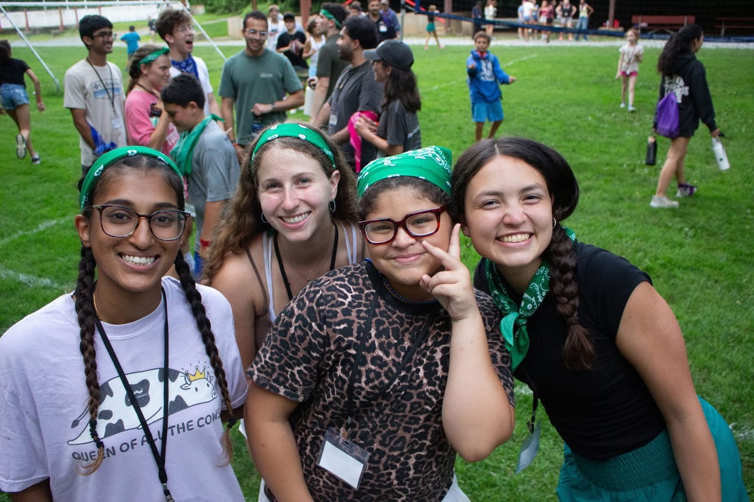 Counselors and a camper in green Kesem bandanas crowded together on the field, grinning at the camera during an afternoon activity.