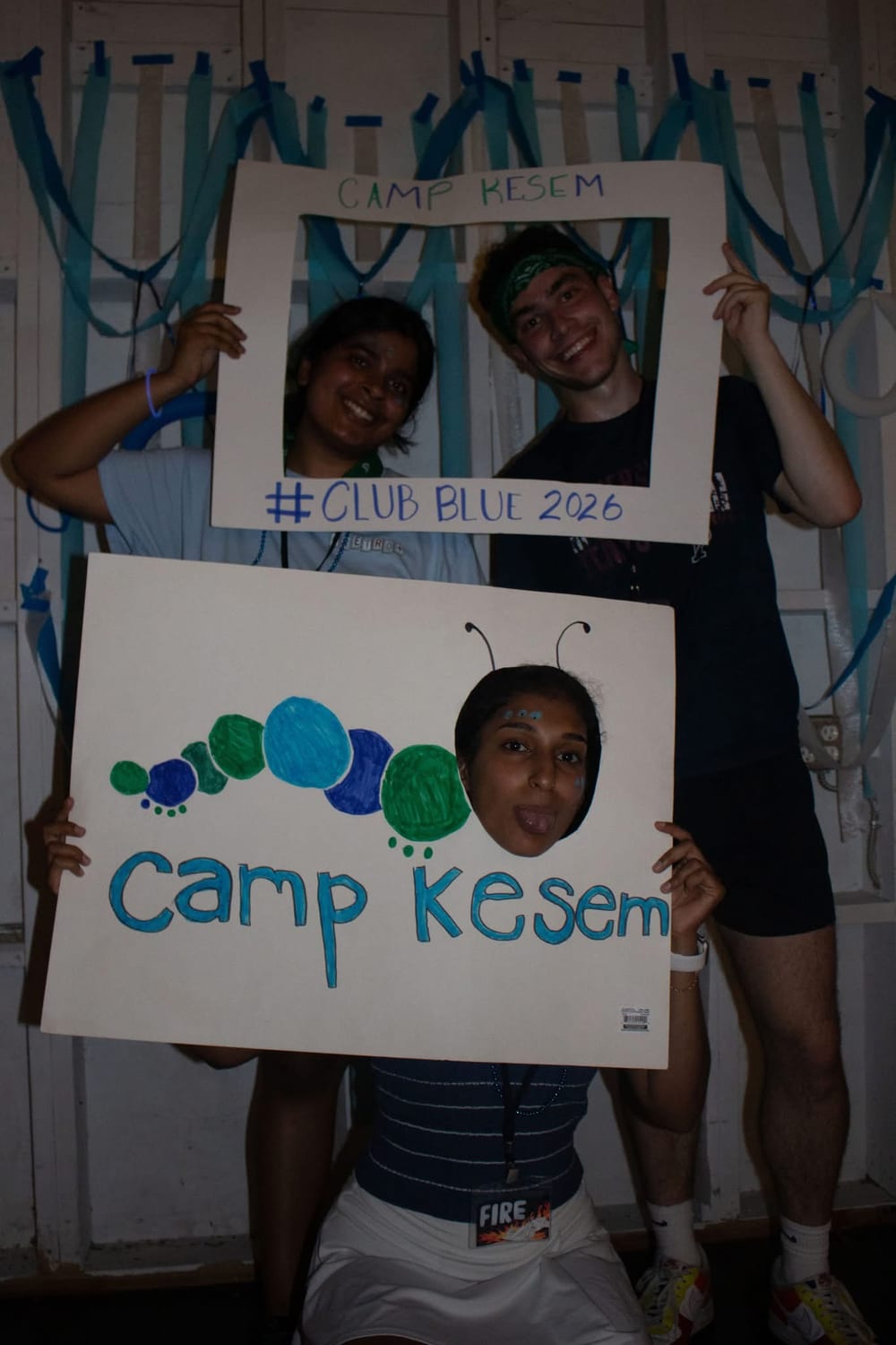 Counselors at an evening event holding a hand-painted "Camp Kesem" caterpillar sign and a homemade photo frame reading "#CLUB BLUE 2026".