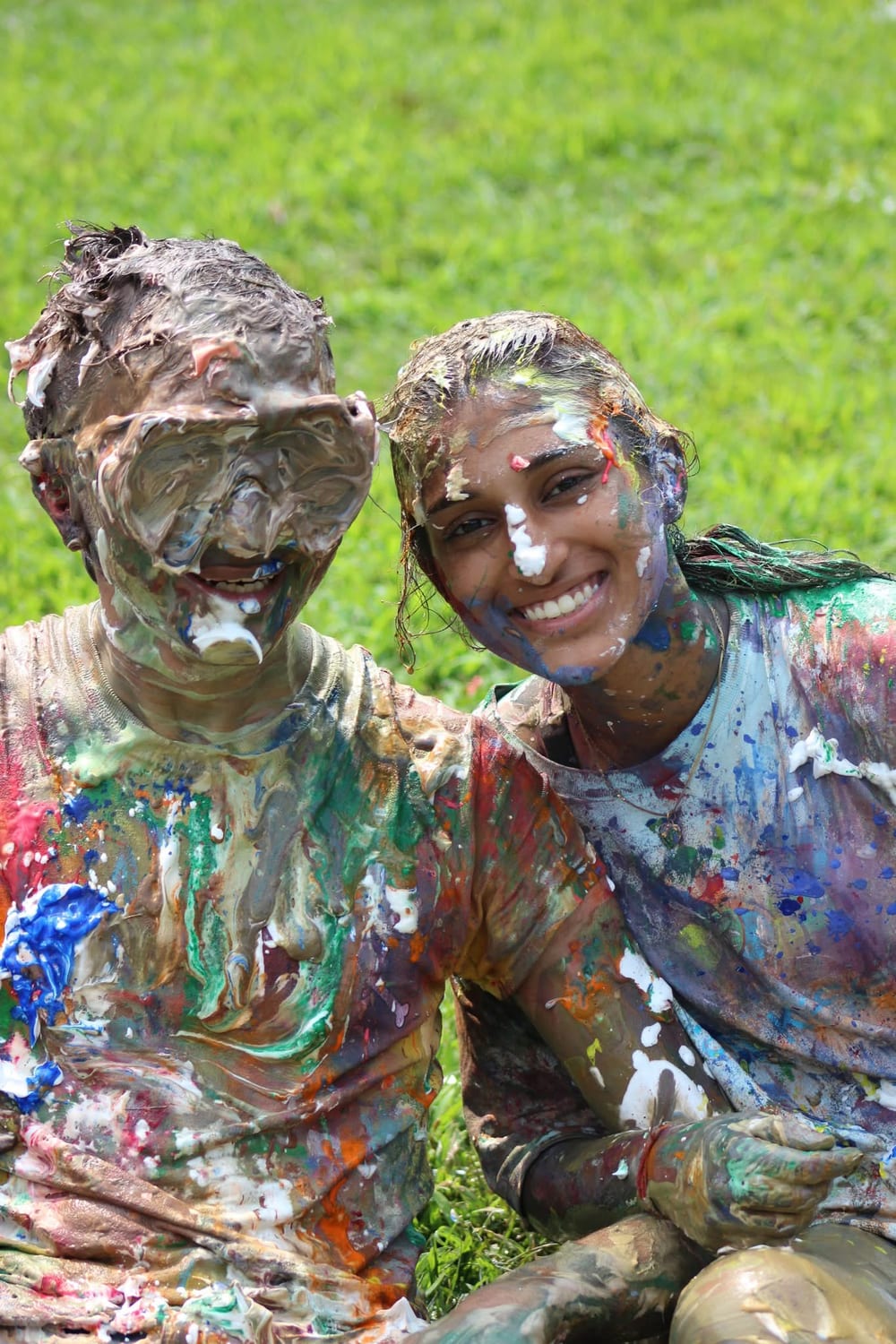 Ava sitting on the grass beside another counselor after the messy games, both covered head to toe in shaving cream and paint, laughing.