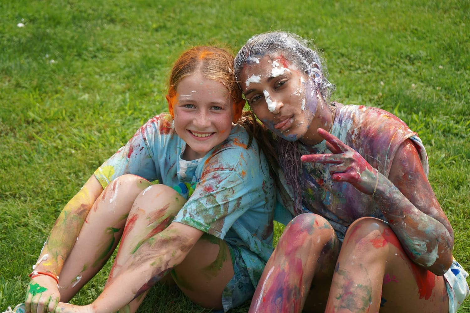 Ava and a camper sitting together on the lawn after the colour games, both painted head to toe, throwing a peace sign.