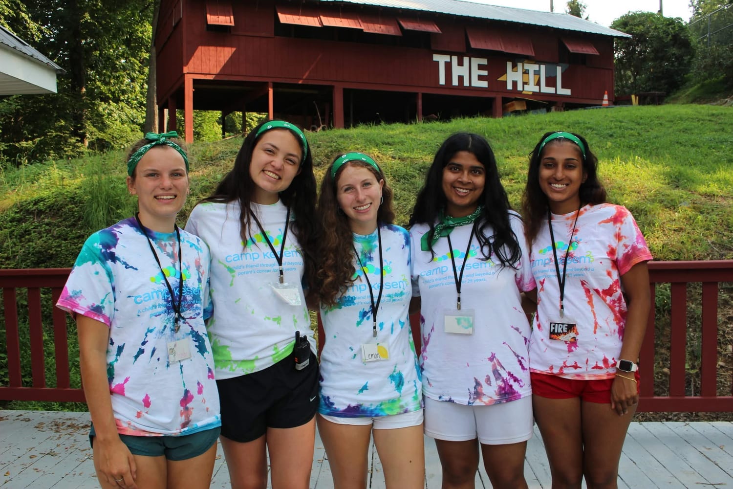 Five Kesem counselors in tie-dyed Camp Kesem shirts and green bandanas, arm in arm on the deck below the red "The Hill" lodge.