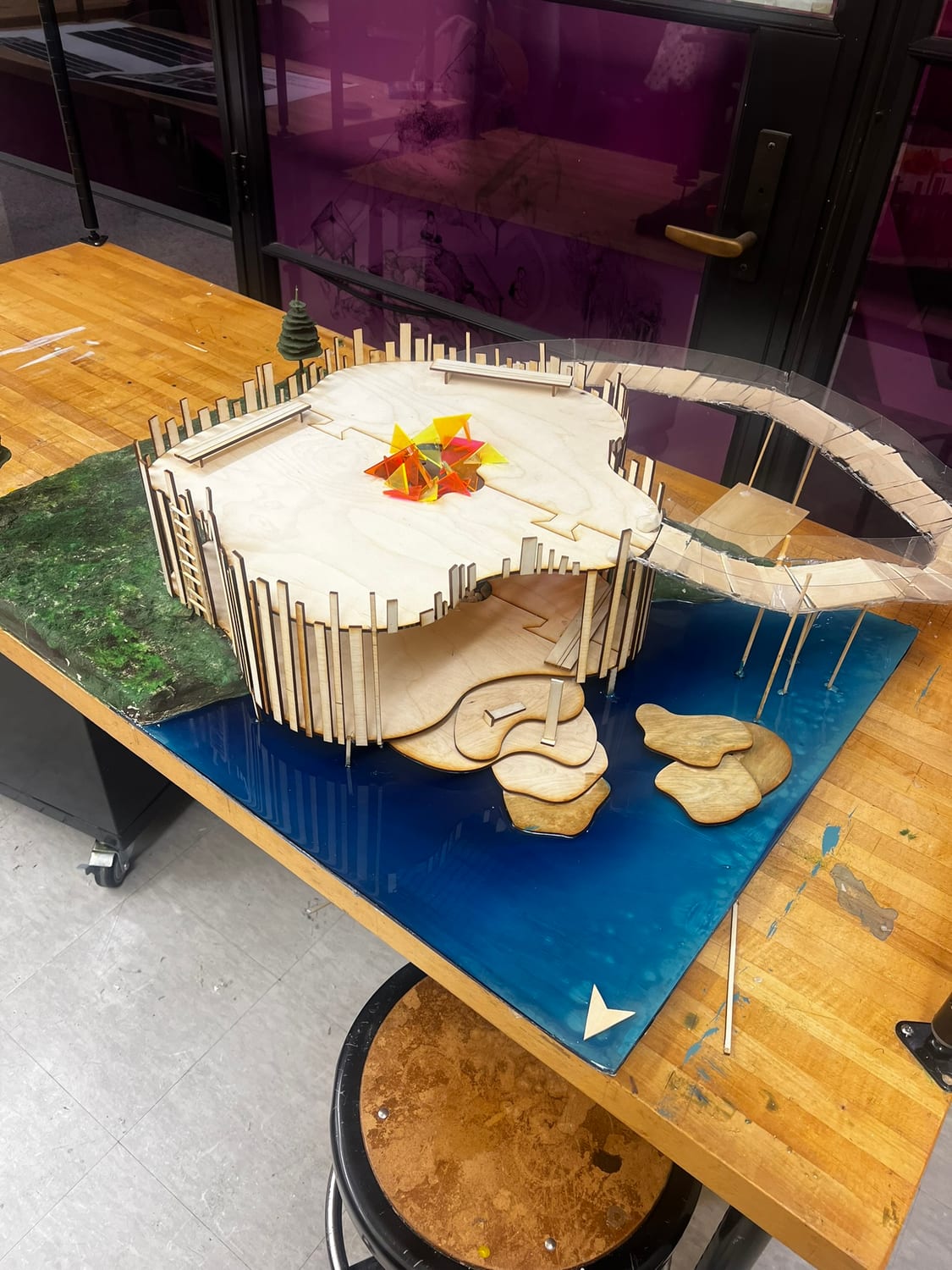 The finished model on the studio workbench: slatted pavilion, poured acrylic river, wooden stepping stones, and the stained-glass fixture at the centre.