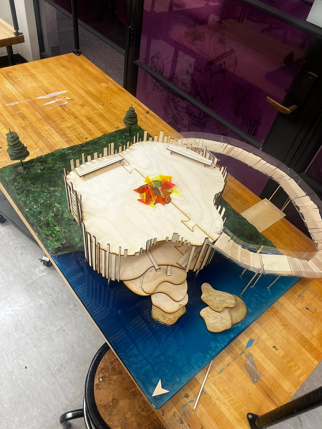 High oblique view of the model — the plan, the walkway, the river base with stepping stones, and conifers on the green terrain.