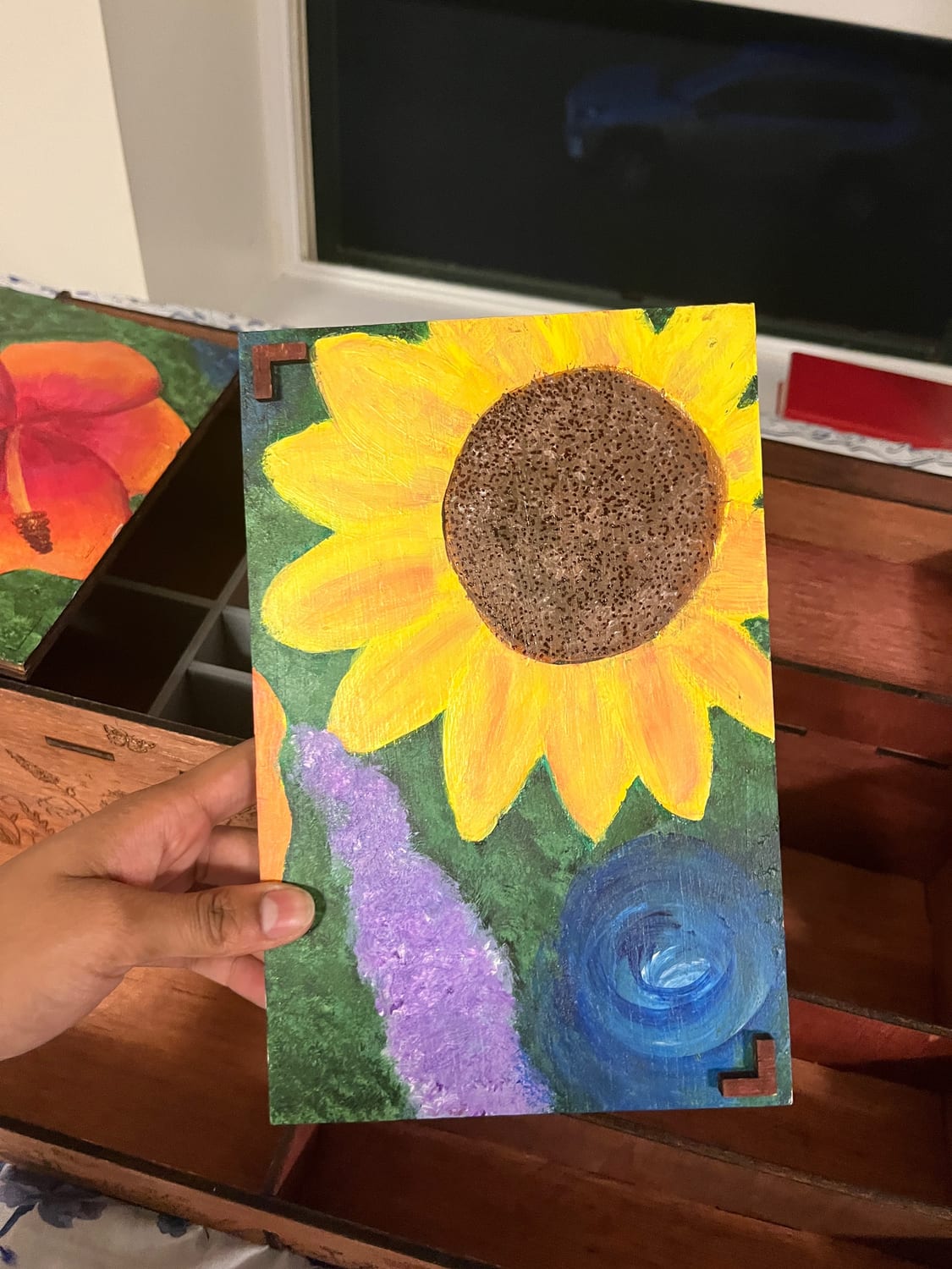 One of the two hand-painted lid panels held in hand — a sunflower over a lavender field and blue swirl — with finger-joint tabs cut along the edges to slot into the case.