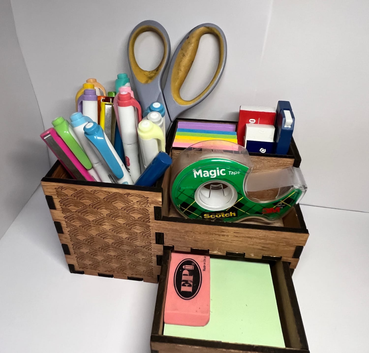In use on a desk — pens and highlighters in the tall bay, a tape dispenser and sticky notes in the tray, an eraser and note pad in the open drawer.