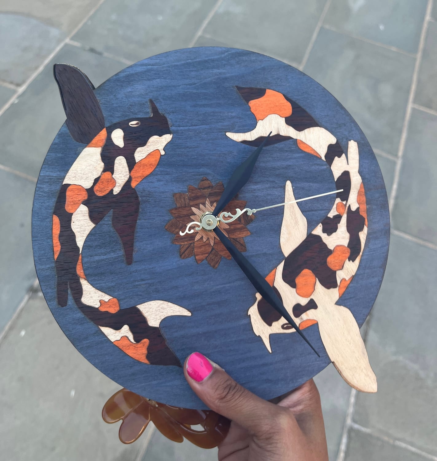The finished koi clock: two calico koi in orange, cream and dark-brown veneer chasing each other in a ring around a turned wooden lotus hub, set into a pond-blue stained ground.