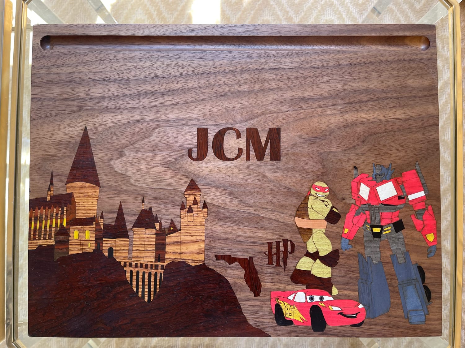 The finished lap desk top: a marquetry scene in a dozen veneers — Hogwarts castle on a dark cliff, the state of Florida, the Harry Potter logo, Raphael, Lightning McQueen and Optimus Prime — with JCM inlaid in the walnut sky above.