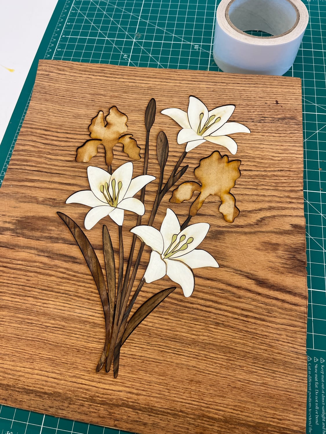 The assembled bouquet — white lily petals with yellow stamens, walnut stems and leaves, iris shapes in a darker wood — laid into the oak ground.