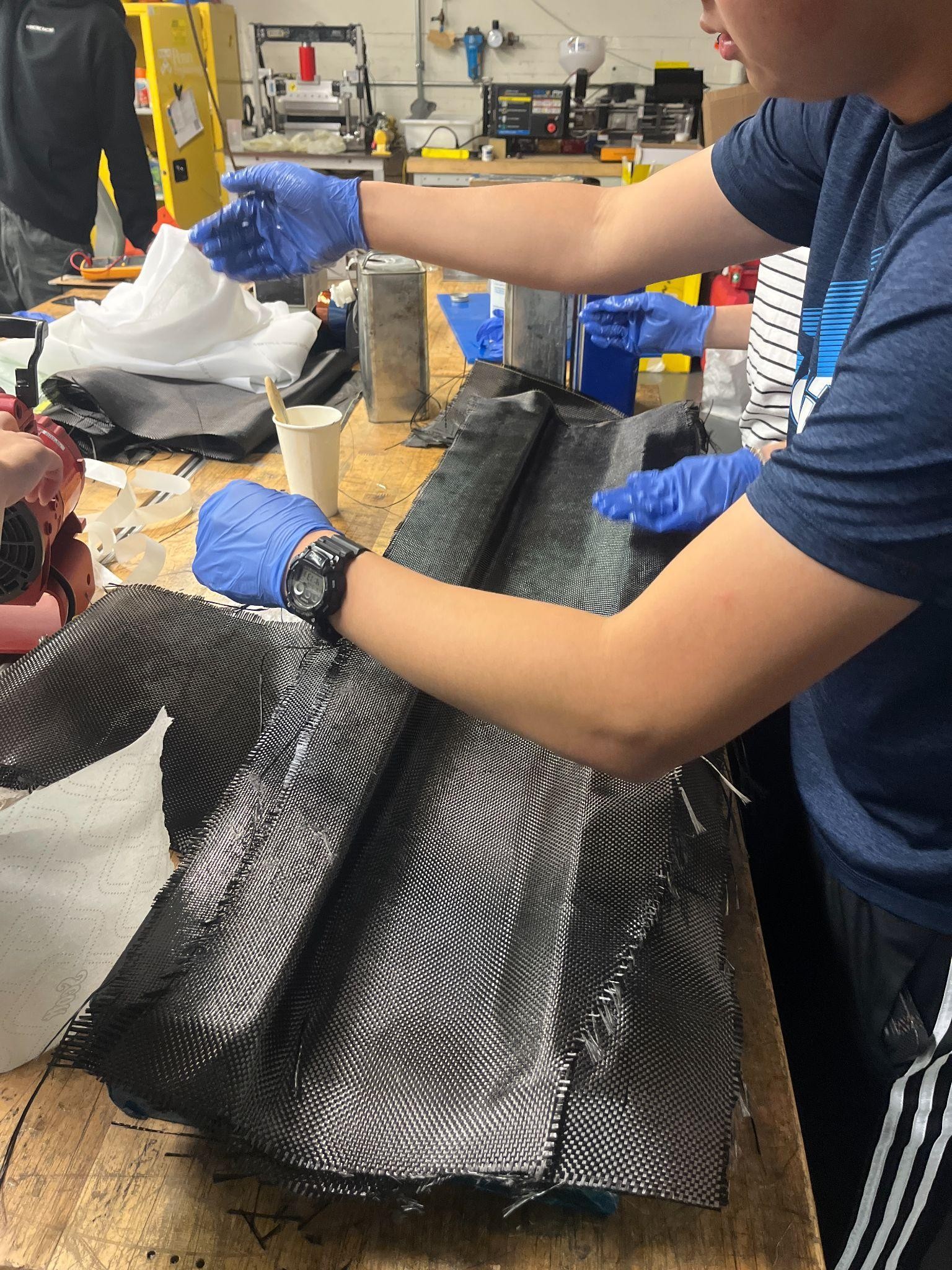 A teammate in nitrile gloves draping a sheet of woven carbon-fiber fabric over the airfoil mold during hand layup, on a cluttered composites bench in the shop.