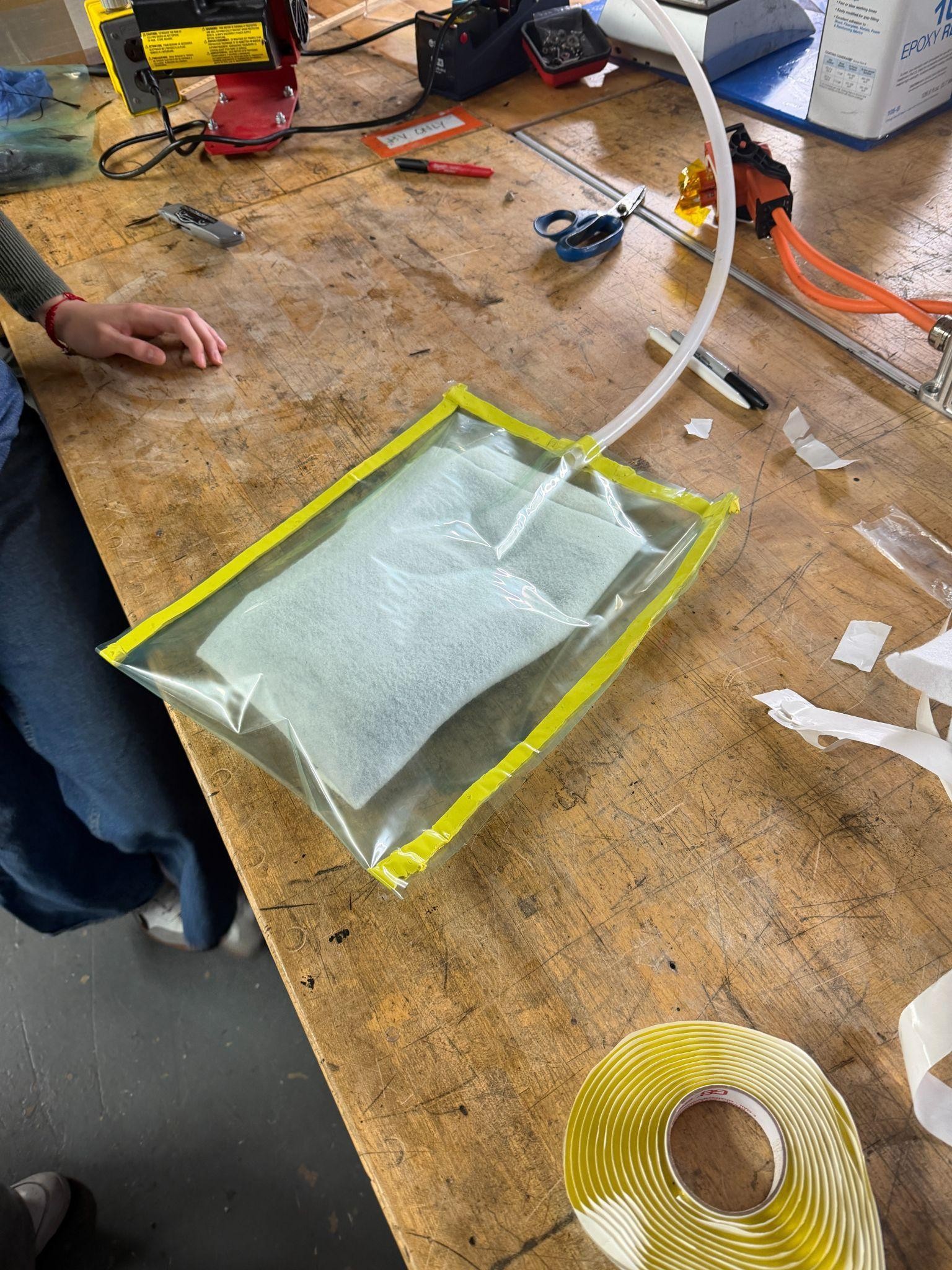 The laid-up part sealed under clear vacuum-bagging film with yellow sealant tape around the perimeter and a vacuum line running in, breather fabric visible through the bag.