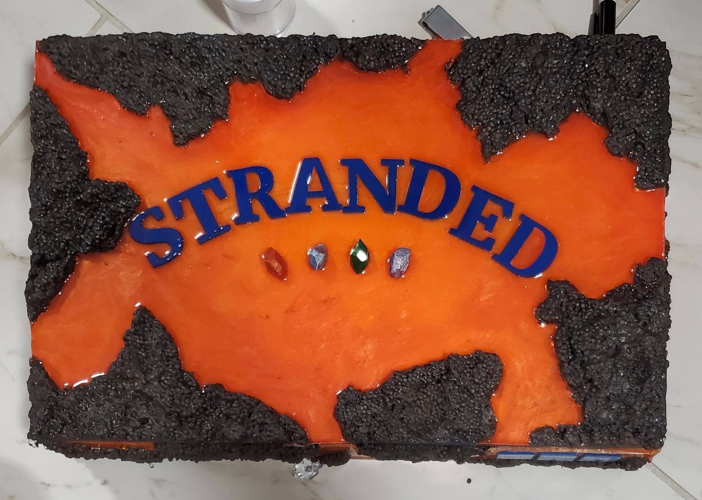 The Stranded box lid — an orange-and-red lava resin surface framed by rough black 'lava rock' edges, the title STRANDED in blue with four inset gemstones.