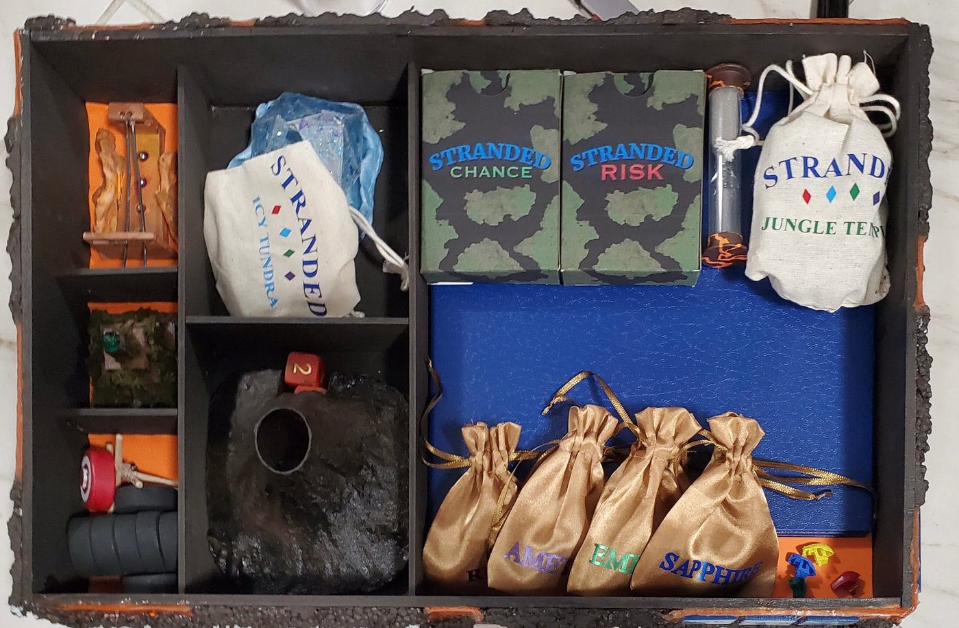 The open box interior — compartments holding the black volcano dice piece with a red die, gold gemstone pouches (Sapphire, Emerald, Amethyst), the Risk and Chance card decks, and the Icy Tundra and Jungle Temple bags.