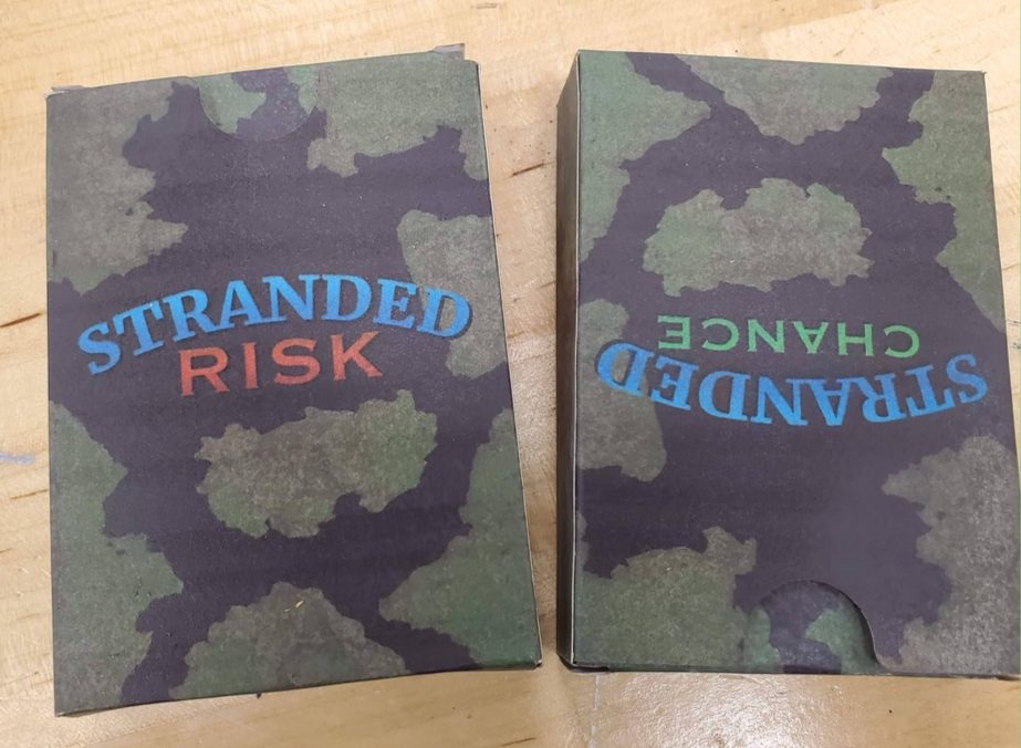 The two custom decks in their boxes — Stranded Risk and Stranded Chance — printed with a green-and-black camouflage pattern.